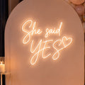 A romantic event backdrop setup featuring a large arched white board with a glowing neon sign that reads 'She said YES'. The board is flanked by a balloon garland in blush pink, cream, and metallic gold, with multiple glass candle holders and lit candles arranged on the wooden floor.