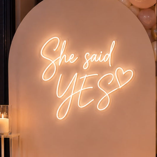 A romantic event backdrop setup featuring a large arched white board with a glowing neon sign that reads 'She said YES'. The board is flanked by a balloon garland in blush pink, cream, and metallic gold, with multiple glass candle holders and lit candles arranged on the wooden floor.
